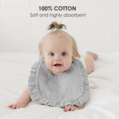 Smiling baby wearing a light gray muslin cotton bib with ruffled edges, lying on a white surface. Text overlay reads '100% COTTON - Soft and highly absorbent.'