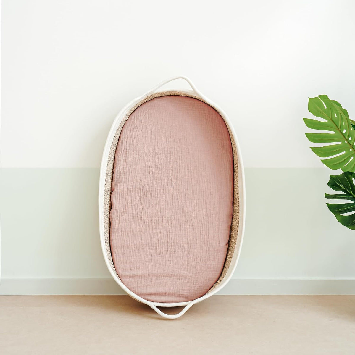 Oval bassinet with a dusty pink fitted muslin sheet lining the interior, shown leaning upright against a two-tone white and sage green wall. The basket has a natural woven texture with white rope handles. A monstera leaf is visible at right.