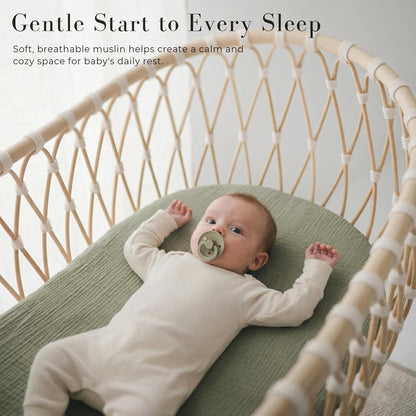 Baby lying on a sage green muslin fitted sheet inside a woven rattan bassinet. The textured cotton sheet has a subtle grid pattern. Text reads 'Gentle Start to Every Sleep' with a breathable muslin callout.