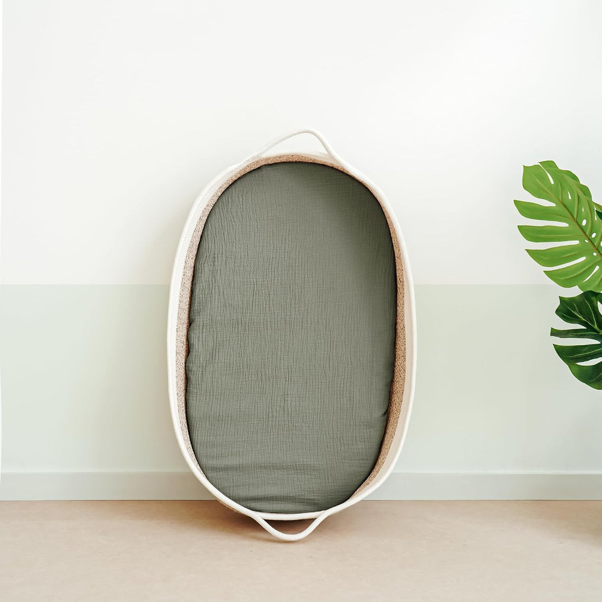 Oval bassinet with a fitted sage green muslin sheet stretched over its mattress, leaning upright against a two-tone white and pale green wall. The bassinet has a natural woven texture and small handles at each end.