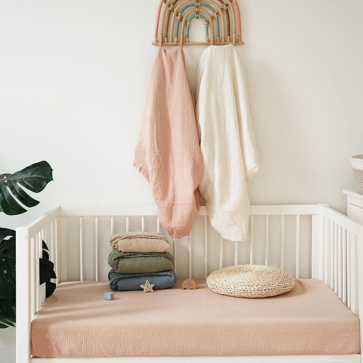 White baby crib with a dusty pink muslin fitted sheet, topped with a stack of folded muslin blankets in blush, olive green, and slate blue. A woven round cushion sits nearby. Two muslin cloths hang above on a rainbow wall hook.