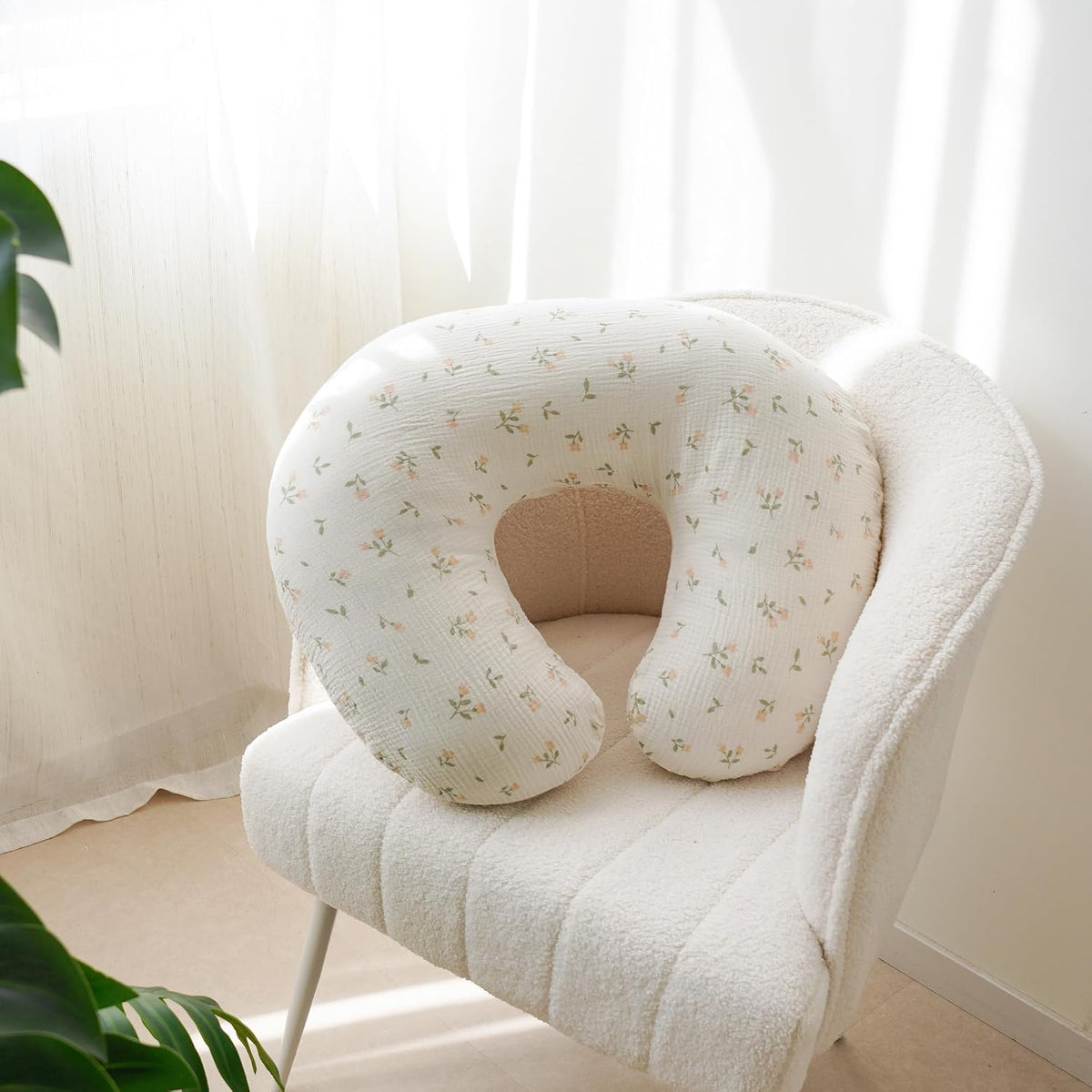U-shaped nursing pillow with a white muslin cover featuring a small floral print in soft pink and green, resting on a cream boucle armchair near a bright window.