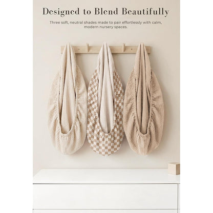 Three baby changing pad covers hanging on a wooden wall hook rack. Left and right covers are solid warm beige muslin; center cover features a tan and cream checkerboard pattern. All have gathered elastic edges.