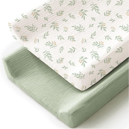 Two changing pad covers stacked at an angle on a white background. The top cover is white with a scattered green leaf and small orange berry print. The bottom cover is solid sage green. Both have a visible textured muslin weave.