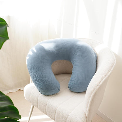 U-shaped nursing pillow cover in soft blue muslin fabric, resting on a white textured armchair. The cover has a slightly crinkled cotton texture and wraps a full nursing pillow, shown in a bright, airy room setting.