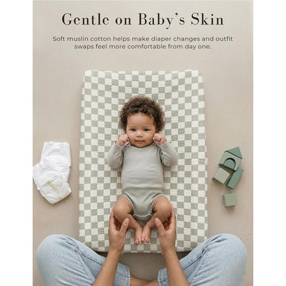Baby lying on a sage green and white checkered muslin changing pad cover. Text reads 'Gentle on Baby's Skin' with a note about soft muslin cotton. A diaper and green toy blocks are visible nearby.