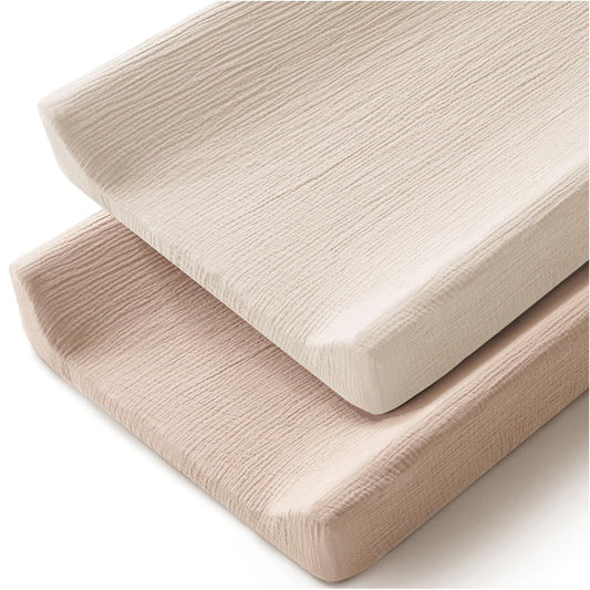 Two changing pad covers stacked at an angle on a white background. The top cover is ivory/cream and the bottom is a warm clay/blush tone. Both feature a crinkled muslin texture with softly rounded corners.