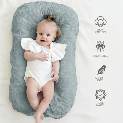 Smiling baby lying in a dusty blue textured cotton baby lounger on a cream surface. Four icons on the right list product features: 100% Cotton, Breathable, Super Soft, and Long Lasting.