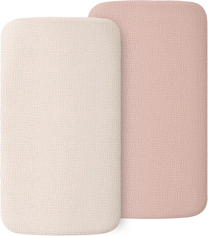 Two rectangular fitted bassinet sheets with rounded corners, shown overlapping. One is off-white/cream and one is dusty pink, both with a visible woven muslin texture.