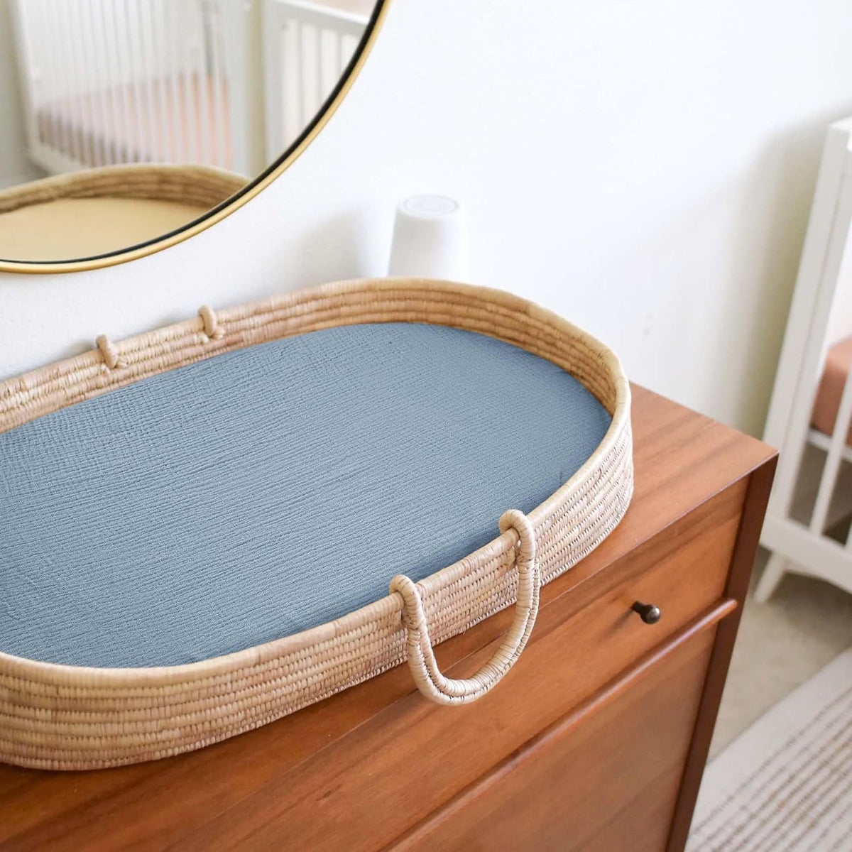 Oval woven rattan changing basket with a fitted dusty blue muslin sheet sitting on a medium-toned wood dresser, with a round gold-framed mirror and white crib visible in the background.