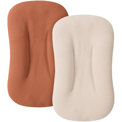 Two fitted baby lounger covers in a textured muslin fabric, shown overlapping. Left cover is terracotta/caramel orange; right cover is cream/off-white. Both have a rounded rectangular shape with contoured edges.