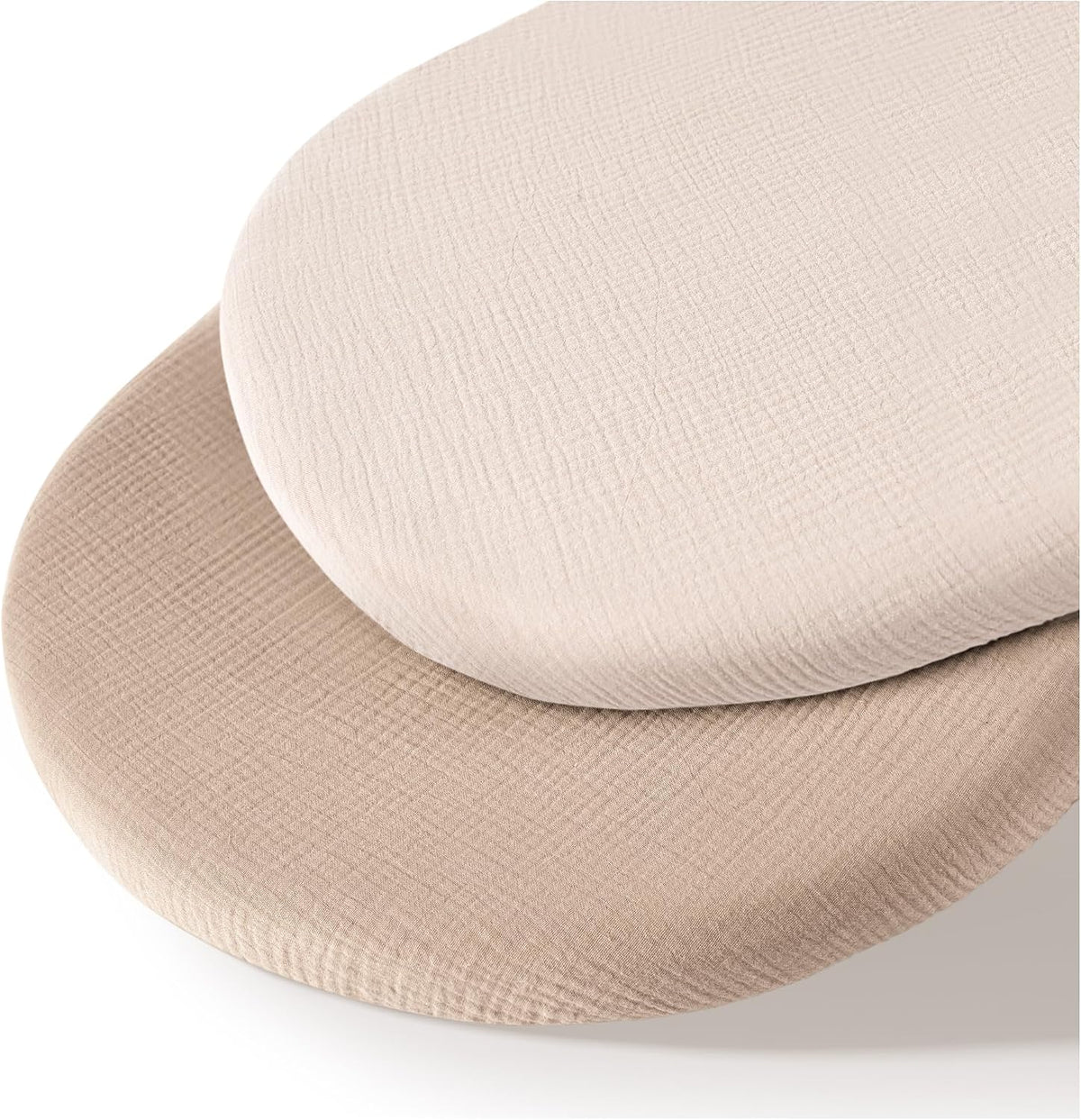 Two oval fitted bassinet sheets stacked at an angle, one cream-white on top and one warm taupe beneath, both showing a soft crinkled muslin texture on a white background.