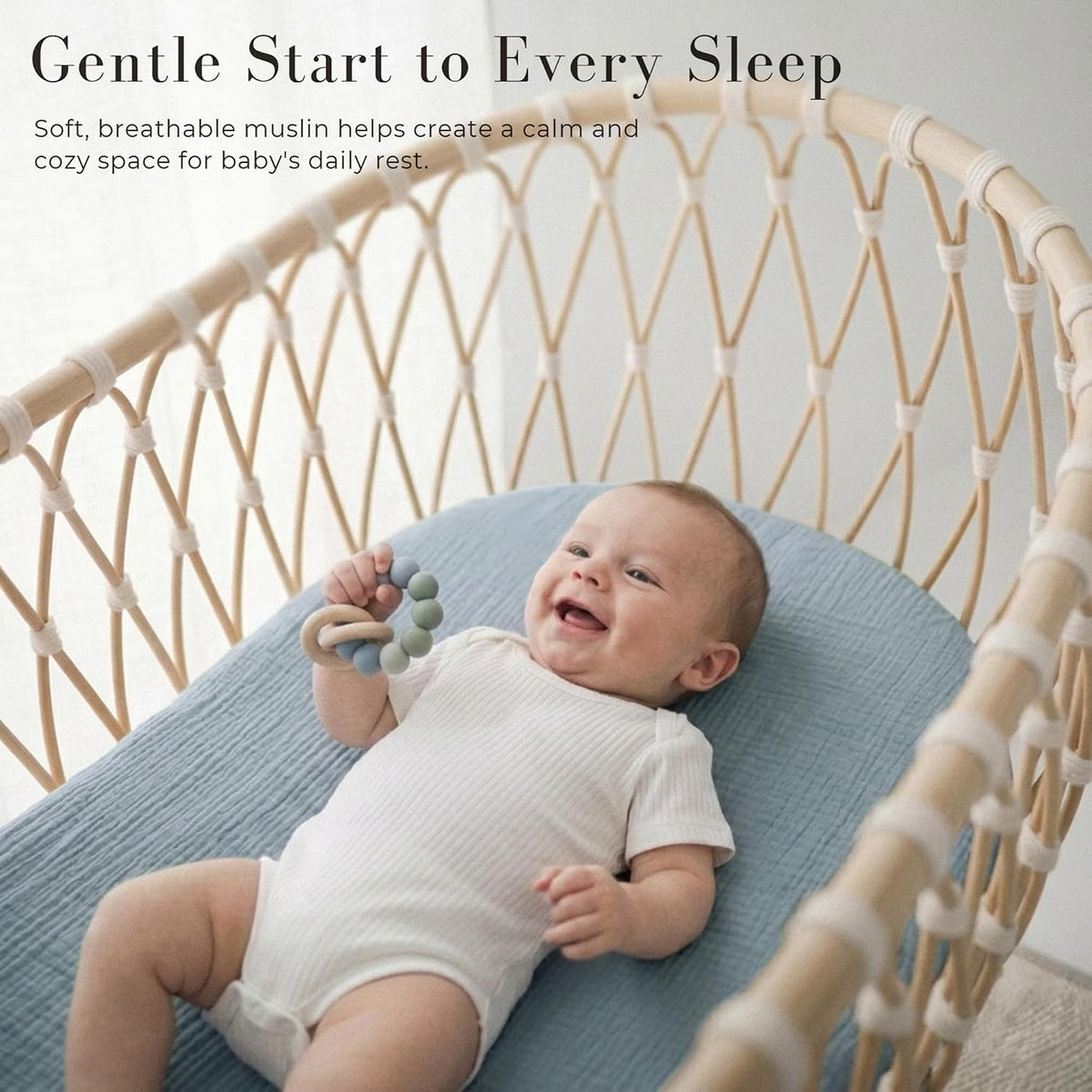 Smiling baby lying on a soft blue muslin fitted sheet inside a woven rattan bassinet, holding a silicone teething ring. Text reads 'Gentle Start to Every Sleep' with a tagline about soft, breathable muslin.