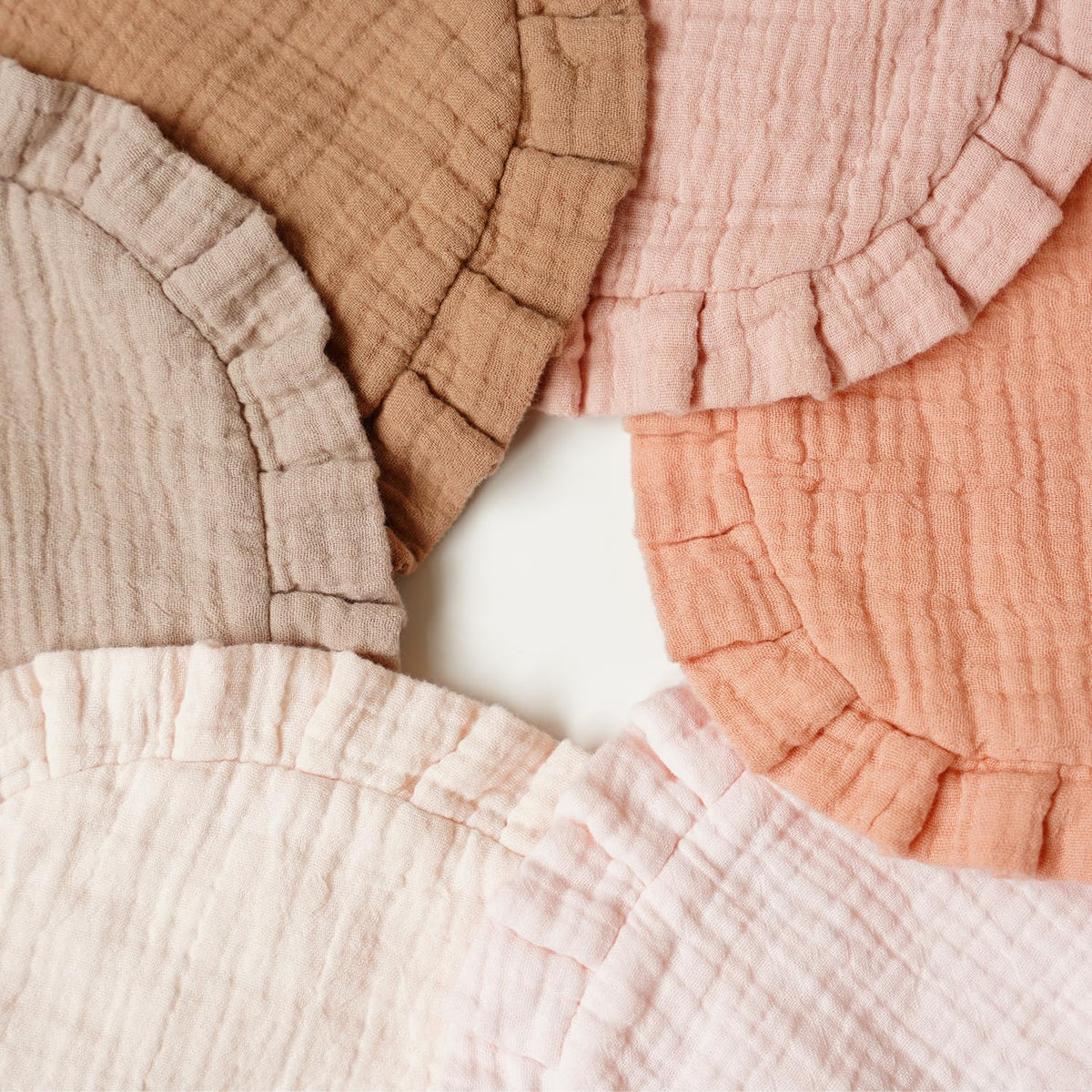 Five muslin cotton baby bibs fanned out in a circle, each with a ruffled edge. Colors include dusty mauve, tan, blush pink, peach-coral, and pale cream, all with a crinkled double-layer texture.