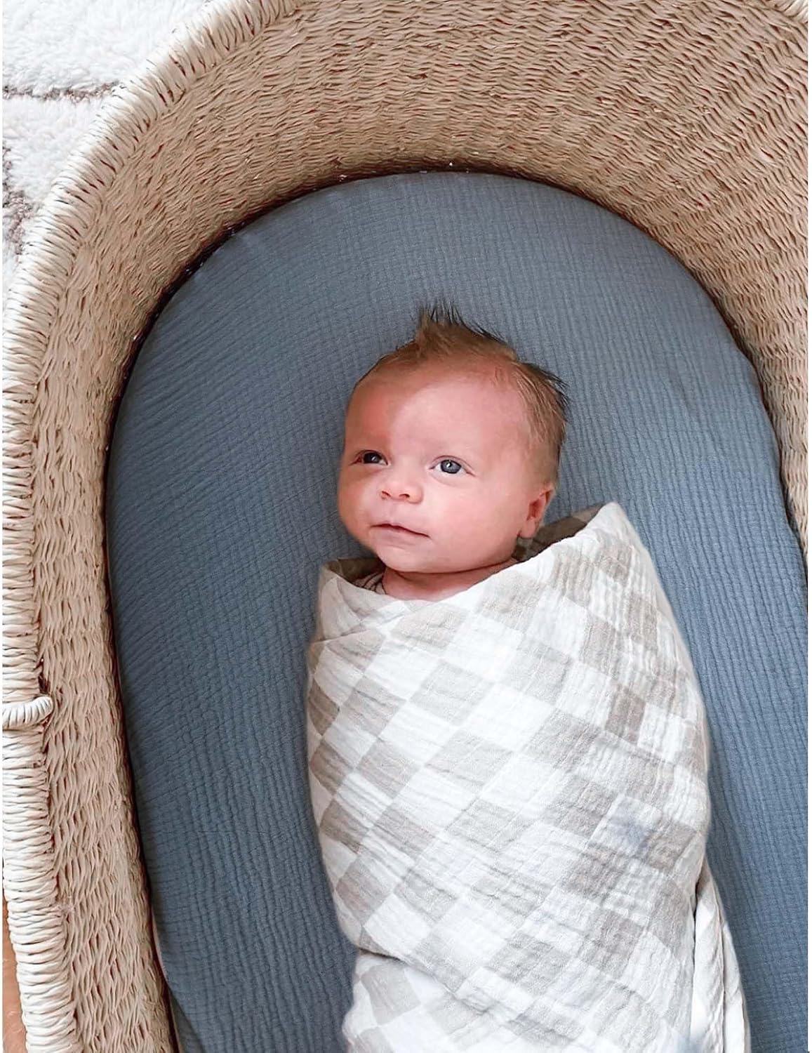 Newborn baby swaddled in a white and tan checkered muslin blanket, lying in a wicker bassinet fitted with a soft slate blue muslin sheet. The textured woven bassinet has a rounded oval shape.