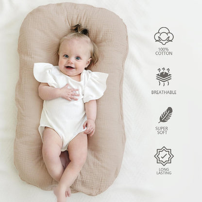 Baby lying on a tan/clay-colored muslin cotton lounger cover, viewed from above. The cover has a soft, textured fabric surface. Icons on the right list: 100% Cotton, Breathable, Super Soft, and Long Lasting.