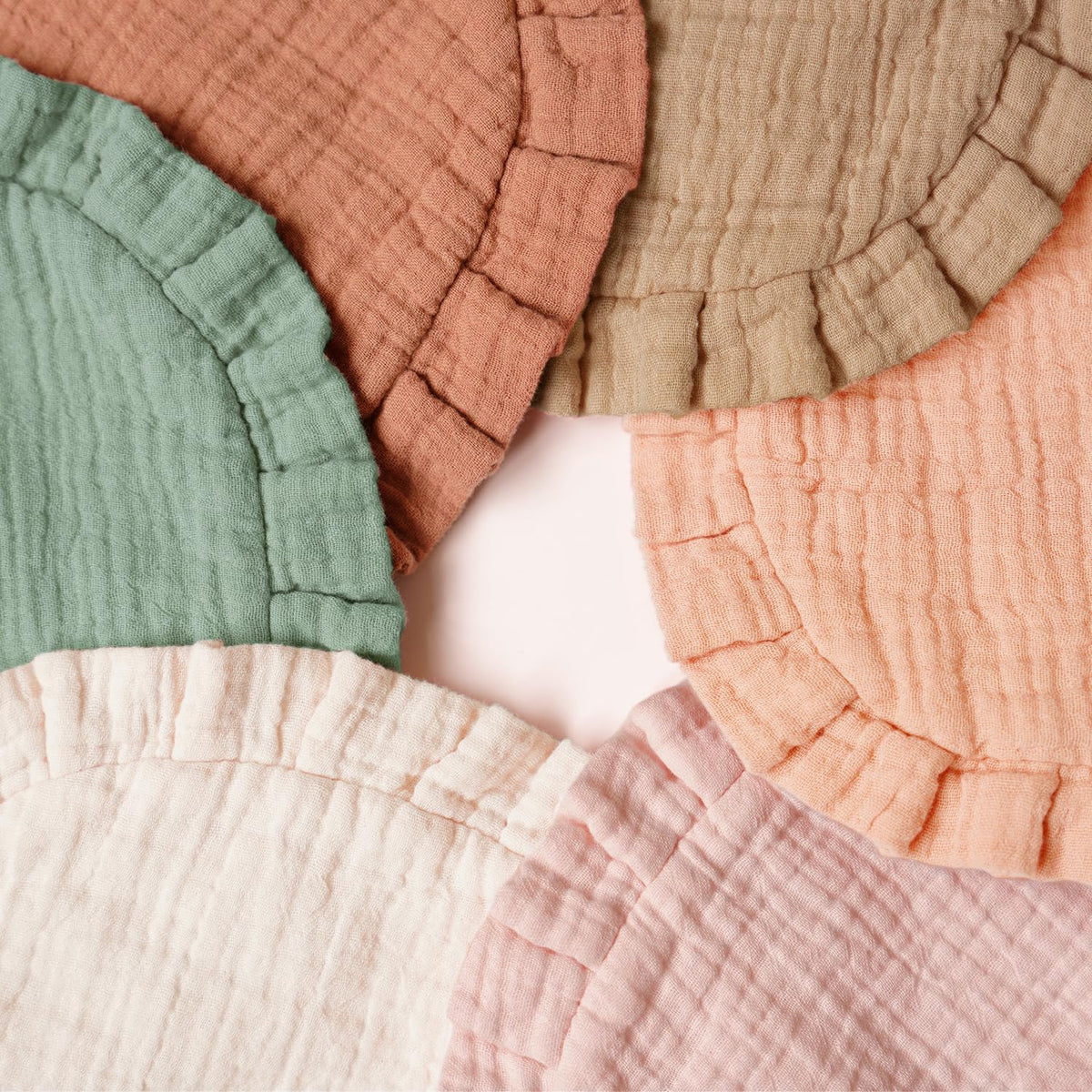 Five round muslin cotton bibs with ruffled edges fanned out in a circle, showing five soft muted colors: sage green, terracotta, olive, peach, and blush pink, with a cream/ivory bib at the bottom center.