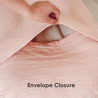 Close-up of a blush pink muslin fabric cover being lifted to reveal an envelope closure opening, with a hand pulling back the textured cotton fabric. Text reads 'Envelope Closure' at the bottom.