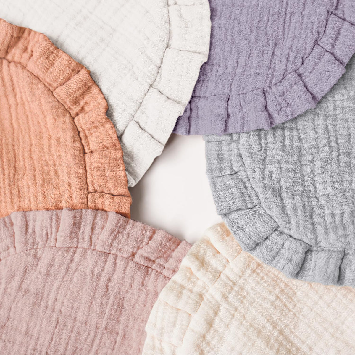 Six round muslin cotton baby bibs fanned out on a white surface, each with a ruffled edge. Colors include terracotta, white, lavender, light gray, dusty pink, and cream.