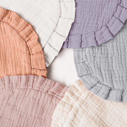 Six round muslin cotton baby bibs fanned out on a white surface, each with a ruffled edge. Colors include terracotta, white, lavender, light gray, dusty pink, and cream.