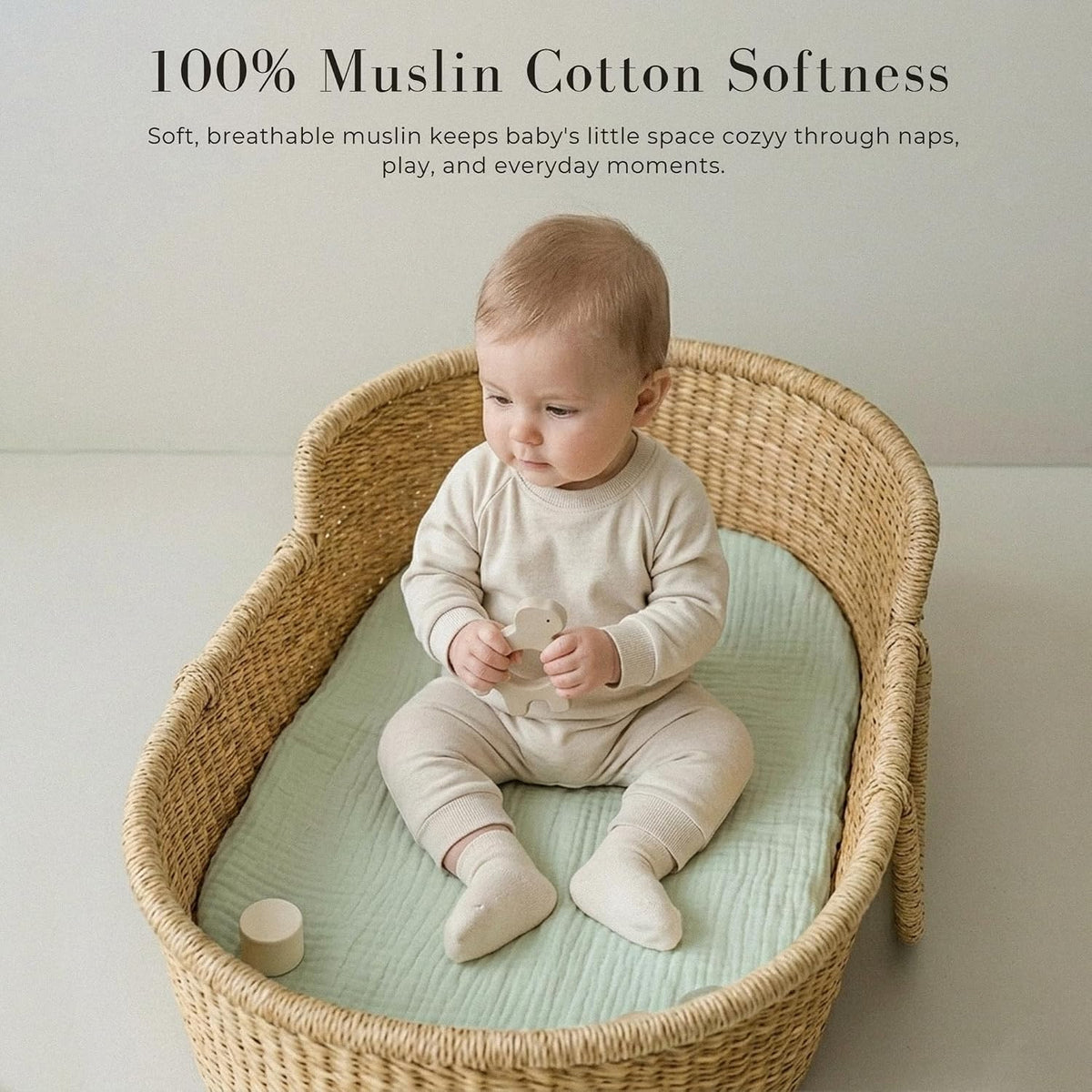 A baby sits in a wicker bassinet fitted with a soft sage green muslin cotton sheet. Text overlay reads '100% Muslin Cotton Softness' with a tagline about breathable comfort for naps and play.