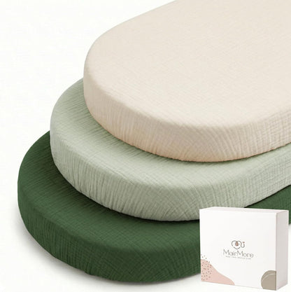 Three oval bassinet fitted sheets stacked together in cream, sage green, and dark forest green, showing a textured muslin fabric surface. A white MairMore branded box is visible in the bottom right corner.