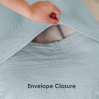 A hand lifts the flap of a light blue textured muslin fabric cover, revealing a white interior lining and the envelope-style opening. Text below reads 'Envelope Closure'.