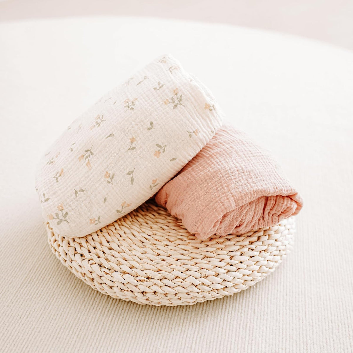 Two folded muslin cotton crib sheets resting on a round woven rattan tray. One sheet is cream with a small floral print in soft pink and green; the other is solid dusty rose. Both show a crinkled muslin texture.