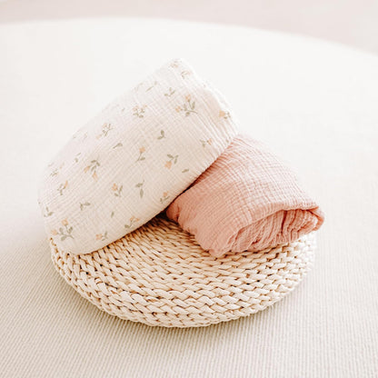 Two folded muslin cotton crib sheets resting on a round woven rattan tray. One sheet is cream with a small floral print in soft pink and green; the other is solid dusty rose. Both show a crinkled muslin texture.