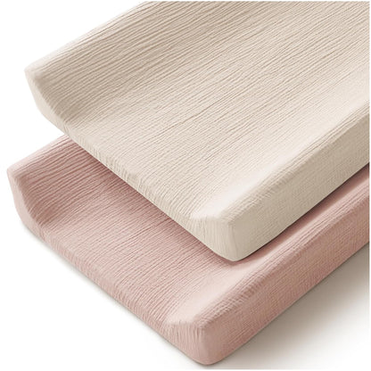 Two changing pad covers stacked at an angle on a white background. The top cover is ivory/cream and the bottom is soft blush pink. Both feature a crinkled muslin cotton texture with fitted edges.