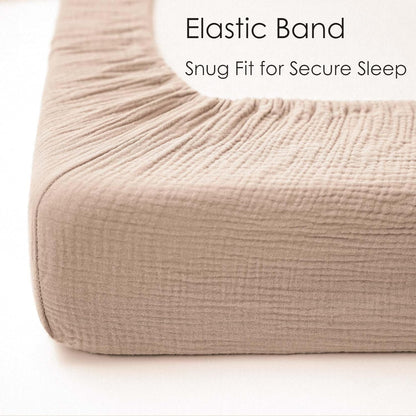 Close-up of a fitted crib sheet in light taupe muslin fabric, showing the elasticated edge stretched over a mattress corner. Text reads 'Elastic Band - Snug Fit for Secure Sleep.'
