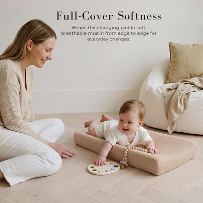 A baby lies on a tan muslin changing pad cover on the floor, smiling and reaching for a colorful toy. A woman sits nearby watching. Text reads 'Full-Cover Softness - Wraps the changing pad in soft, breathable muslin from edge to edge for everyday changes.'