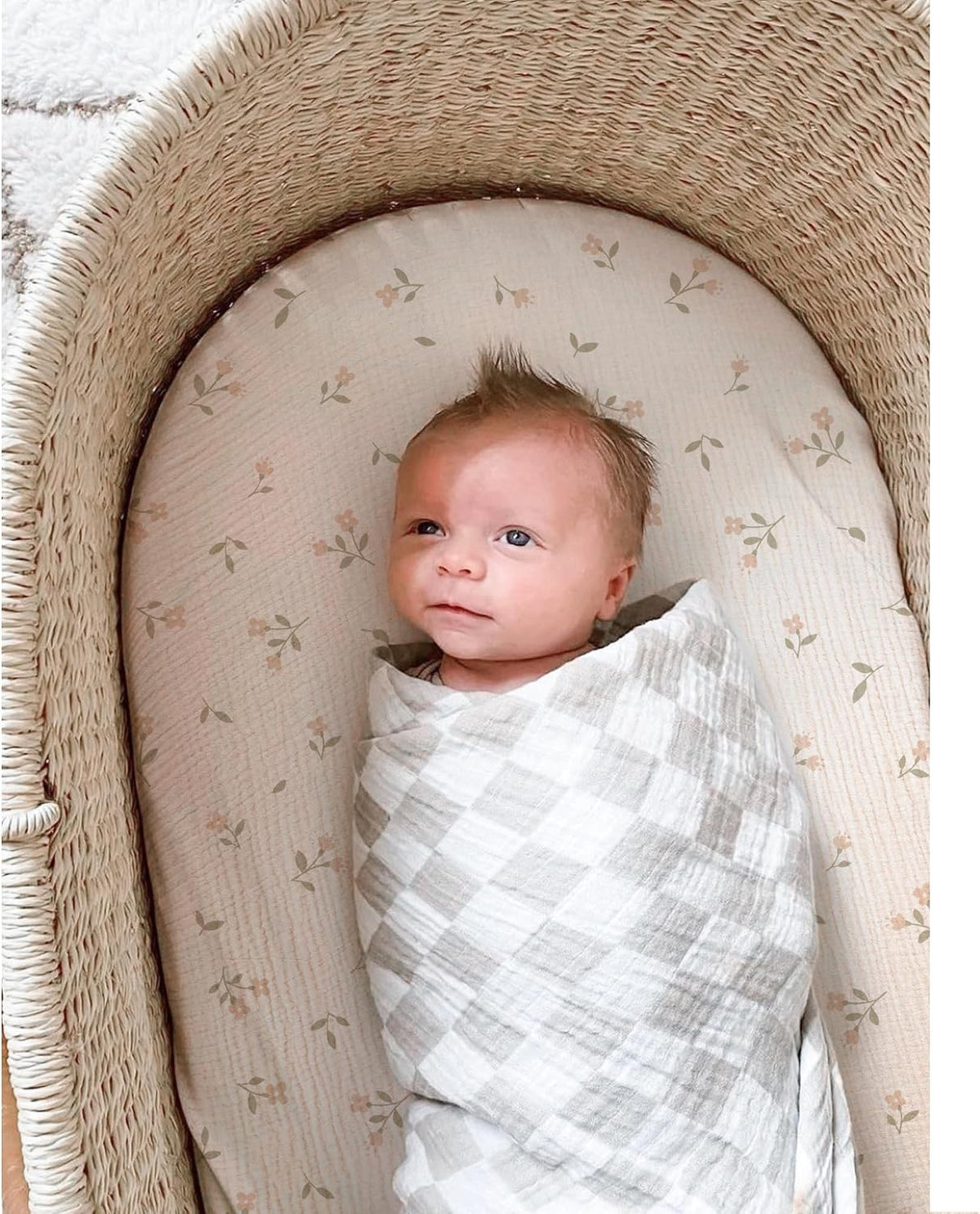 Newborn baby swaddled in a gray and white checkered muslin blanket, lying in a wicker bassinet fitted with a cream cotton sheet featuring a small floral print with pink blooms and green leaves.