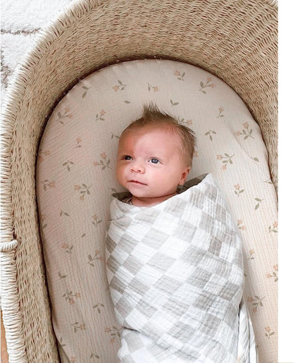 Newborn baby swaddled in a gray and white checkered muslin blanket, lying in a wicker bassinet fitted with a cream cotton sheet featuring a small floral print with pink blooms and green leaves.