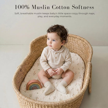 A baby sits in a wicker bassinet fitted with a white muslin sheet featuring a small floral print. Text reads '100% Muslin Cotton Softness' with a tagline about breathable comfort for naps and play.