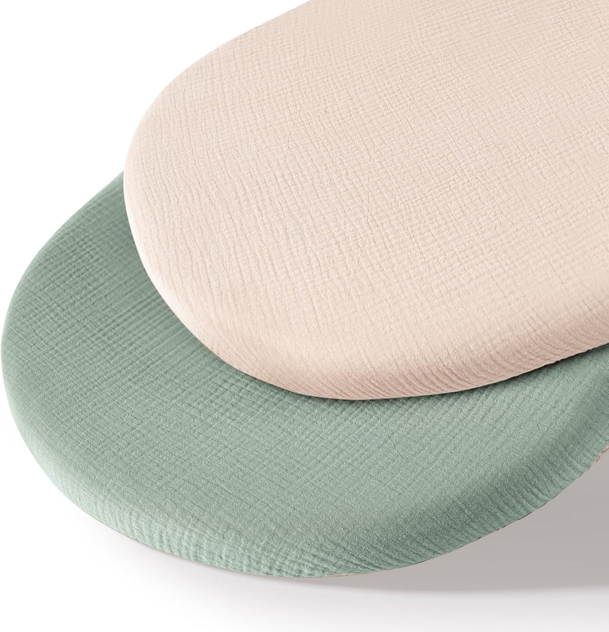 Two oval fitted bassinet sheets stacked together, one in soft blush pink and one in muted sage green, both showing a textured muslin weave fabric on a white background.