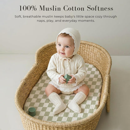 Baby sitting in a wicker bassinet on a green and white checkered muslin fitted sheet. Text reads '100% Muslin Cotton Softness' with tagline about breathable muslin for naps, play, and everyday moments.