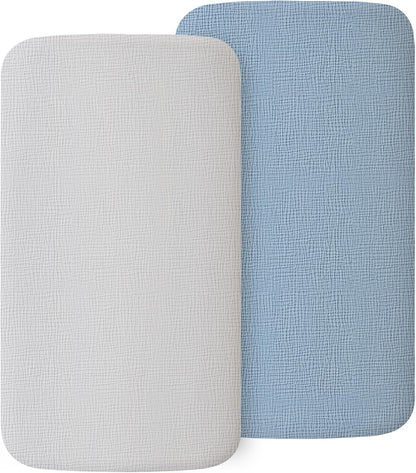 Two rectangular fitted bassinet sheets with rounded corners and a visible muslin weave texture, shown overlapping. One sheet is white, the other is light blue. Both have a soft, breathable woven fabric surface.