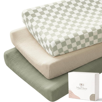 Three stacked changing pad covers in muslin fabric: top cover has a sage green and white checkerboard pattern, middle is solid light beige, bottom is solid sage green. A white MairMore gift box sits in the foreground.