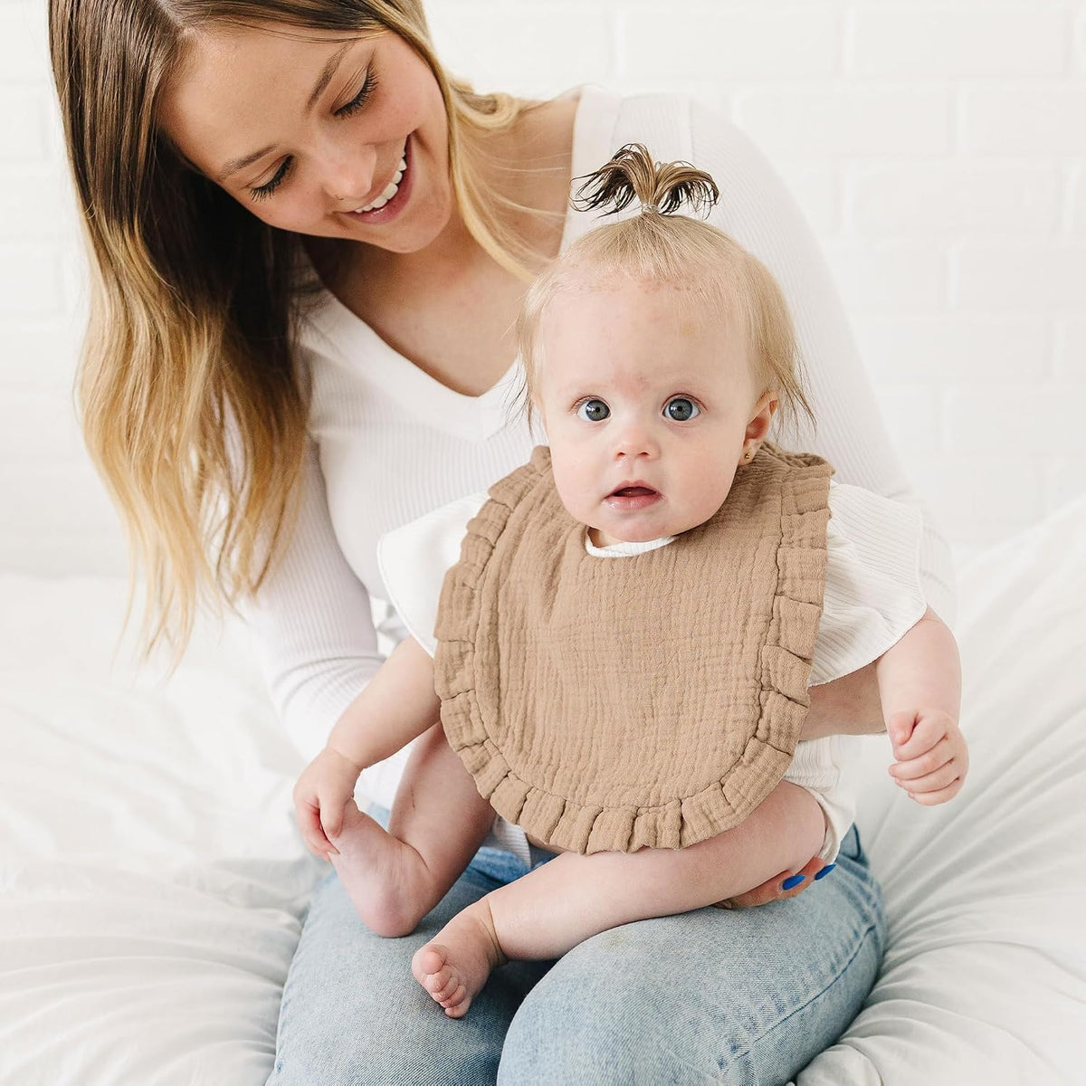 Baby wearing a tan muslin bib with ruffled edges, seated on an adult's lap against a white background. The bib has a textured, gauze-like fabric and decorative ruffle trim around the perimeter.
