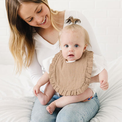 Baby wearing a tan muslin bib with ruffled edges, seated on an adult's lap against a white background. The bib has a textured, gauze-like fabric and decorative ruffle trim around the perimeter.