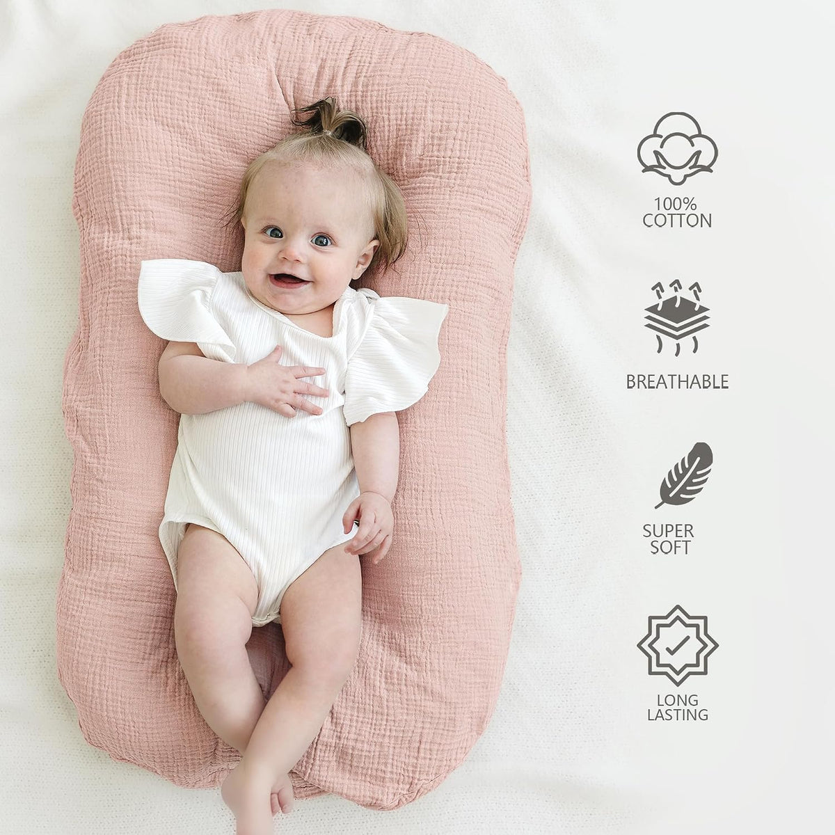 A smiling baby in a white ruffled onesie lies in a dusty pink muslin baby lounger on a cream surface. Icons on the right highlight: 100% Cotton, Breathable, Super Soft, and Long Lasting.