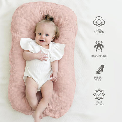 A smiling baby in a white ruffled onesie lies in a dusty pink muslin baby lounger on a cream surface. Icons on the right highlight: 100% Cotton, Breathable, Super Soft, and Long Lasting.