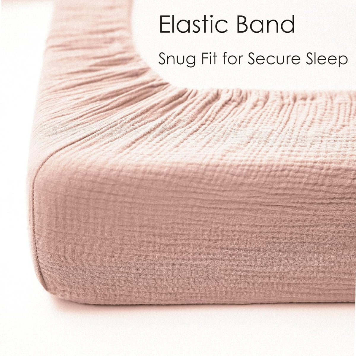 Close-up corner view of a dusty pink muslin cotton fitted crib sheet stretched over a mattress, showing gathered elastic band along the edge. Text reads 'Elastic Band - Snug Fit for Secure Sleep.'