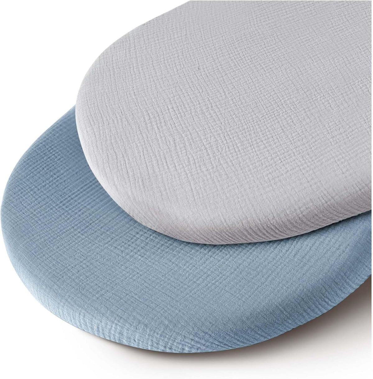 Two oval fitted bassinet sheets stacked at an angle, one light gray on top and one dusty blue beneath, both showing a soft, crinkled muslin cotton texture with fitted edges wrapping around padded mattress forms.
