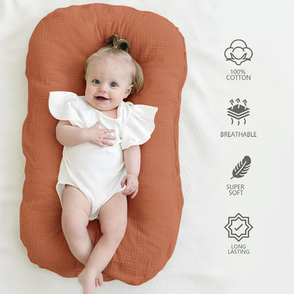 Baby lounger cover in terracotta/caramel-toned textured cotton muslin, shown with a smiling infant lying on it. Right side features four icons with text: 100% Cotton, Breathable, Super Soft, and Long Lasting.