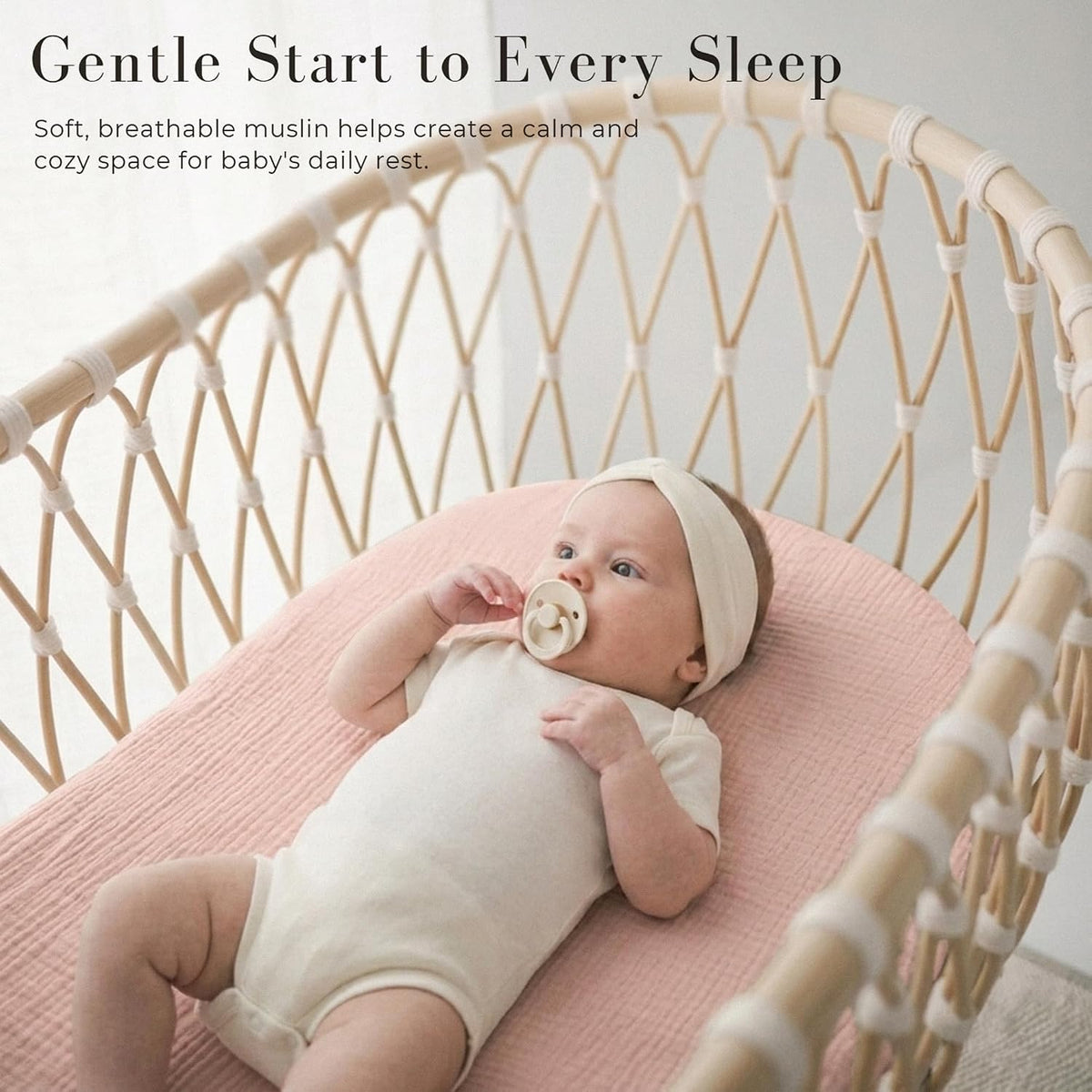 Baby lying on a soft pink muslin fitted sheet in a woven rattan bassinet, wearing a cream onesie and white headband with a pacifier. Text reads 'Gentle Start to Every Sleep' with a breathable muslin callout.