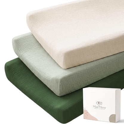 Three stacked changing pad covers in cream, sage green, and dark forest green, showing a crinkled muslin cotton texture. A white MairMore gift box is shown in the lower right corner.
