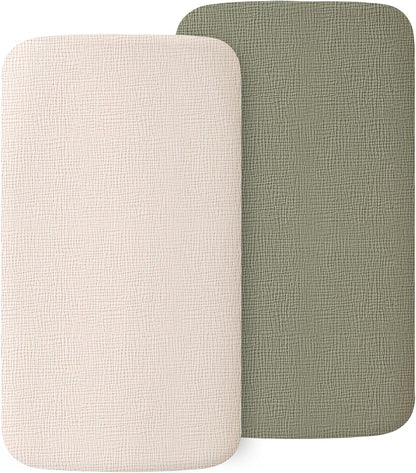 Two rectangular fitted bassinet sheets with rounded corners displayed side by side. Left sheet is cream/off-white, right is muted sage green. Both show a visible woven muslin texture on a white background.