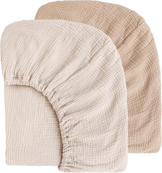 Two muslin fitted crib sheets displayed overlapping on a white background. The front sheet is ivory/off-white and the back sheet is warm taupe/ash. Both show a crinkled muslin texture with elasticized edges.